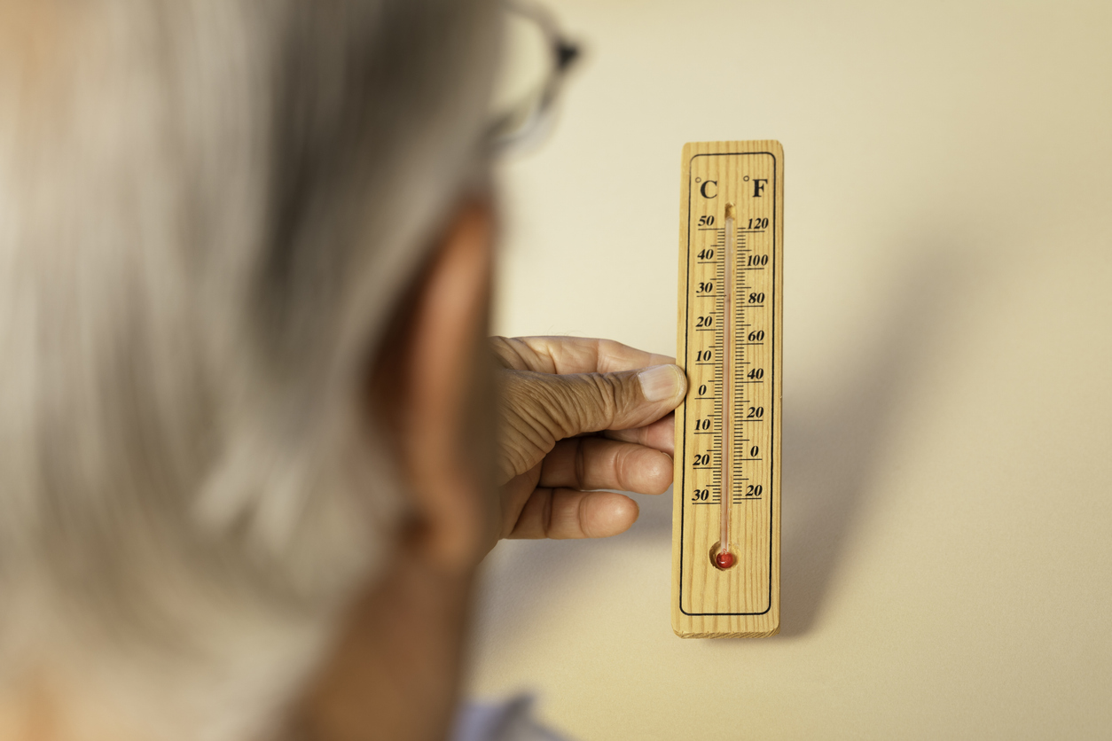Over the shoulder view of senior man holding thermometer in hand showing high temperatures in front of beige background.