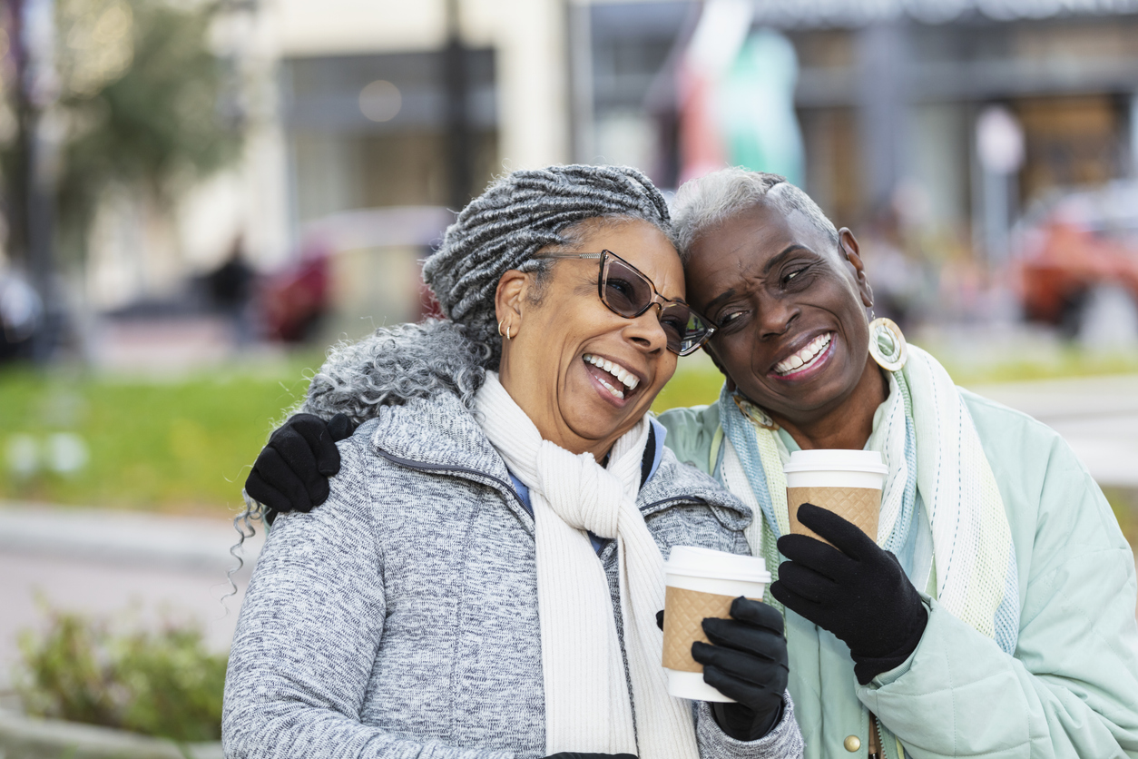 Two senior women sitting outdoors on a city park bench conversing and drinking coffee, smiling and laughing. They are wearing jackets, scarves and gloves to stay warm on a chilly autumn or winter day. The African-American woman wearing eyeglasses is in her 70s. Her friend is in her 60s.