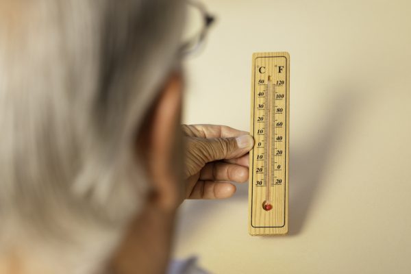 Over the shoulder view of senior man holding thermometer in hand showing high temperatures in front of beige background.