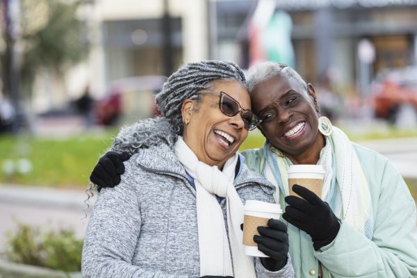 Two senior women sitting outdoors on a city park bench conversing and drinking coffee, smiling and laughing. They are wearing jackets, scarves and gloves to stay warm on a chilly autumn or winter day. The African-American woman wearing eyeglasses is in her 70s. Her friend is in her 60s.