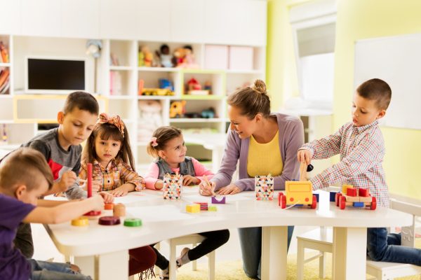 Happy teacher sketching and talking to a smiling little girl at kindergarten while other children are playing with educational  toys.